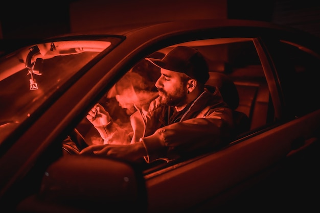 Photo man in a cap driving car smoking at night in a garage lit with a red light, sports car