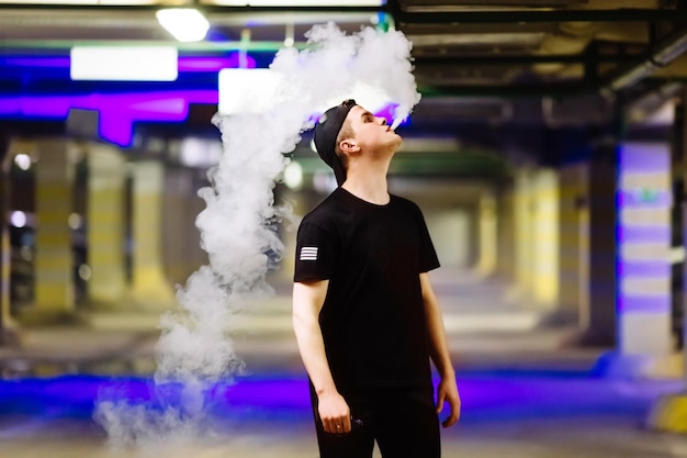 모자를 쓴 남자가 전자 담배를 피우고 증기 구름을 방출합니다.