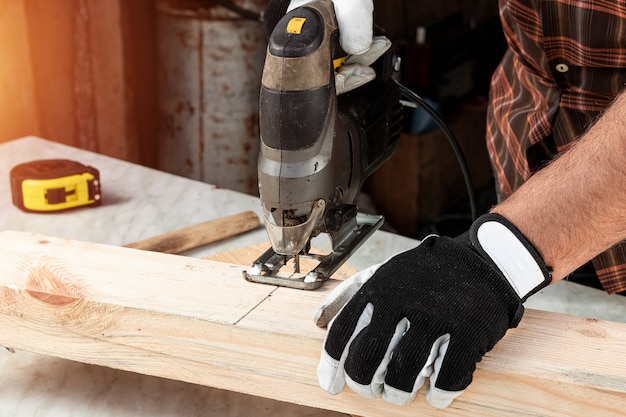 A man carpenter cuts a wooden beam using an electric jigsaw, male hands with an electric jigsaw closeup.