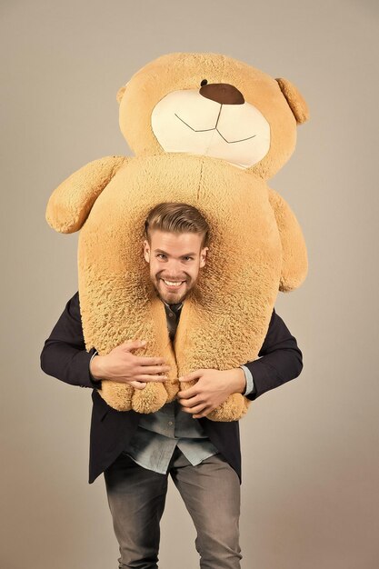 Foto l'uomo porta l'orsacchiotto gigante sul collo sfondo grigio concetto di regalo di compleanno orsacchiotto peluche piacevole sorpresa ragazzo felice faccia barbuta tiene giocattolo orsacchiotto uomo vestito formale carino regalo sorpresa