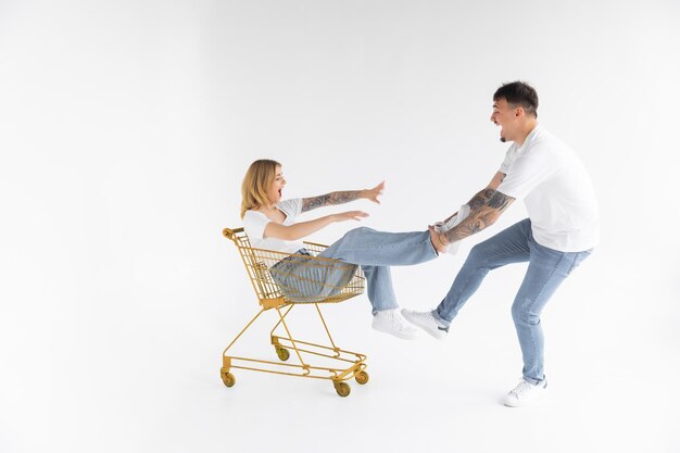 Man carries a woman in a shopping cart on a white background Cheerful young couple