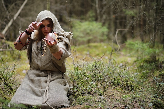 A man in a cassock spends a ritual in a dark forest with a crystal ball and book