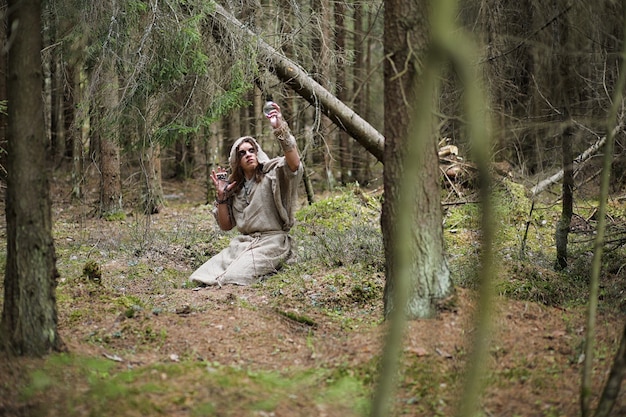 A man in a cassock spends a ritual in a dark forest with a crystal ball and book