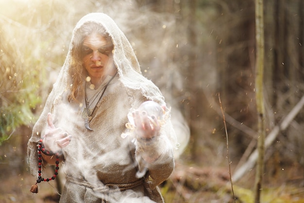 A man in a cassock spends a ritual in a dark forest with a crystal ball and book