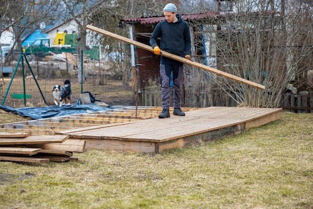 man in casual attire meticulously carrying lumber for backyard deck construction with a playful dog in the background