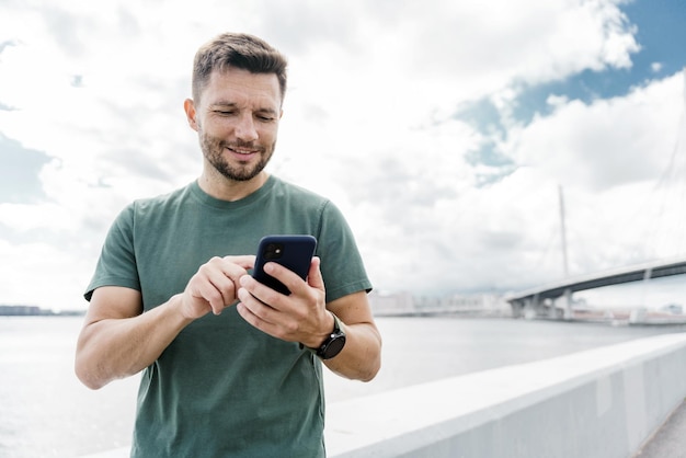 Мужчина в повседневной одежде с помощью смартфона на набережной и небесном фоне, воплощающем городскую связь