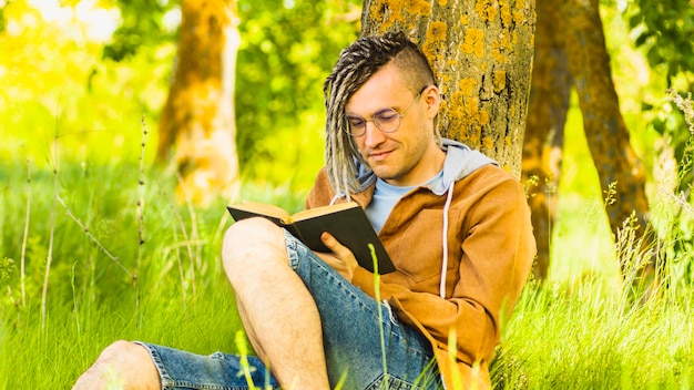 A man in casual clothes and glasses reads literature under a tree A hipster with dreadlocks is relaxing with a book in the woods Male with a book is sitting on the grass on a clear summer day