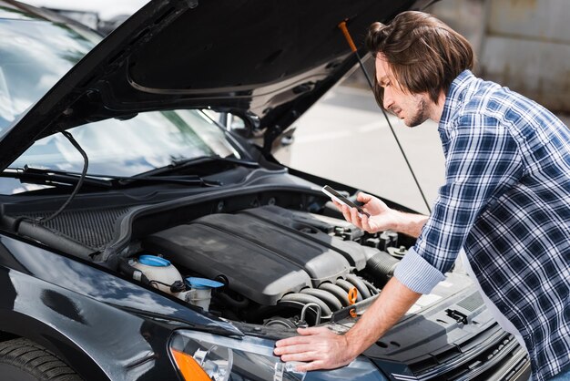 Man in casual clothes holding smartphone in hand and standing near broken auto with open trunk car