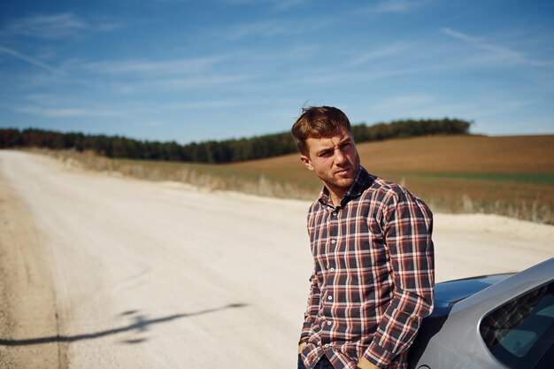 Man in casual clothes leaning on his car outdoors on the road. Rural scene.