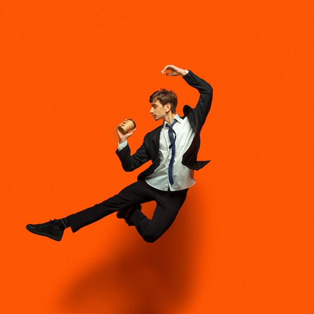 Man in casual office style clothes jumping and dancing isolated on bright orange background. business, start-up, open-space, inspiration concept. flexible ballet dance. hurrying up, drinking coffee