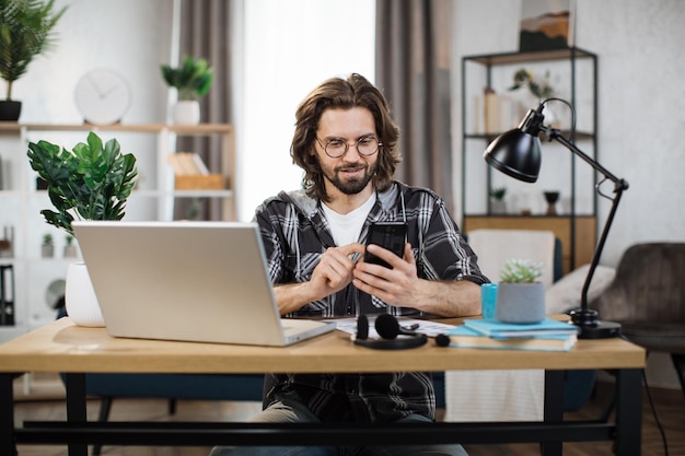Man in casual outfit typing on mobile phone while sitting at workplace with laptop
