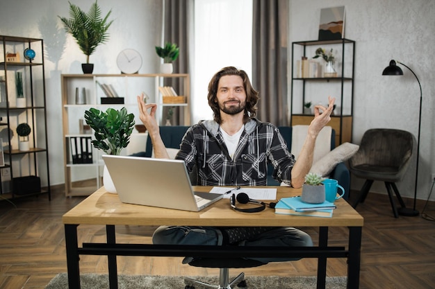 Man in casual wear sitting on workplace with modern laptop and meditating with opened eyes