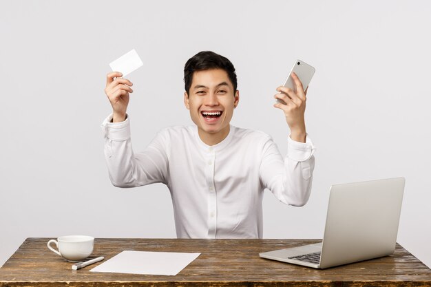 Man celebrating, hooray yes gesture, made good deal, ordered by best price. Asian businessman raising hands up, holding smartphone and credit card, smiling joyfully, read good news laptop display