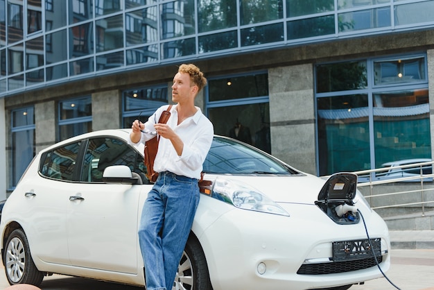 Uomo che carica la sua auto elettrica di lusso alla stazione all'aperto di fronte ai nuovi edifici moderni della città