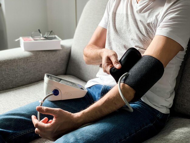 Foto uomo che controlla la pressione sanguigna a casa seduto su un divano grigio problema stagionale organizzando il monitor a mano