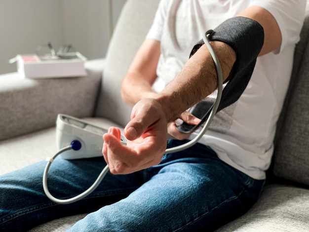 Photo man checking blood pressure at home sitting on gray sofa seasonal issue arranging monitor on hand