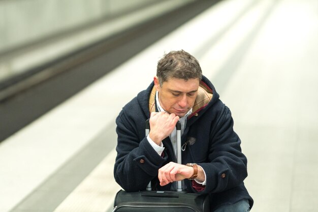 写真 鉄道駅のプラットフォームで荷物と一緒に座っている間に時間をチェックする男性