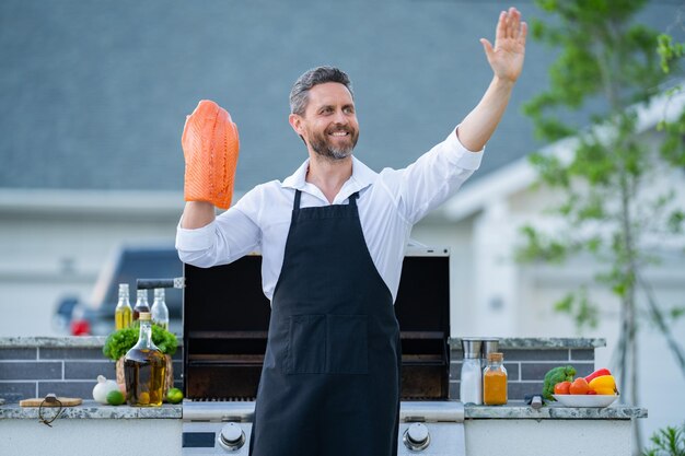 Foto l'uomo in grembiule da chef tiene il pesce salmone alla griglia del barbecue cuoco maschio che prepara barbecue all'aperto pesce barbecue