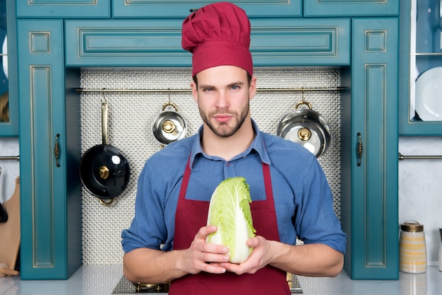 Man chef in red hat, apron hold chinese cabbage vegetable in kitchen. Vegetarian, health, diet concept. Food preparation, cooking, culinary, cuisine.