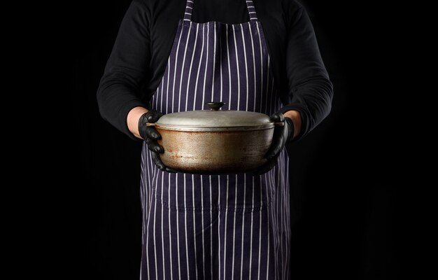 Photo man chef in a striped apron holds an aluminum cauldron with a lid, black background