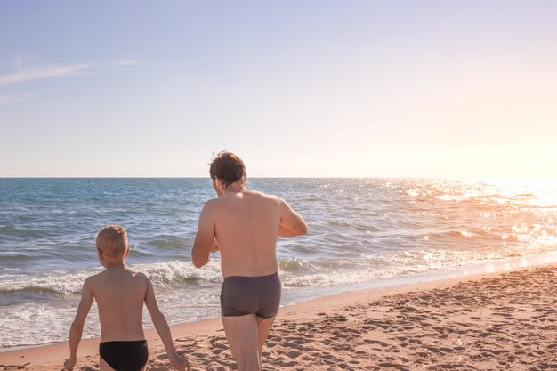 明るい日差しに照らされて、男と子が砂浜を歩いて海へ。夏休みとレクリエーション、旅行と観光。スペースをコピーします。