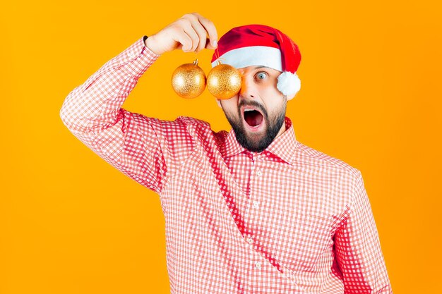 Мужчина в новогодней шапке Деда Мороза держит перед глазами новогодние игрушки, шары золотистого цвета и кричит.