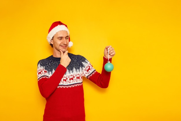 Man in christmas sweater with reindeer red santa claus hat stands with new year tree toy in his hand