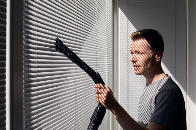 Man cleaning dust from window blind by vacuum cleaner