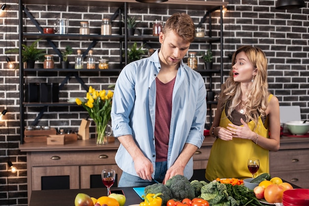 Man cooking. Blonde-haired appealing stylish girlfriend coming to the kitchen and seeing her man cooking