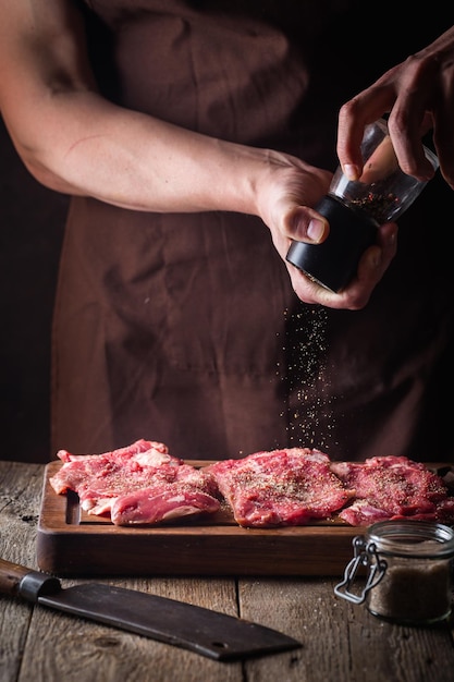 キッチンで肉ステーキを調理する男。木製の背景にシェフの塩とコショウの肉