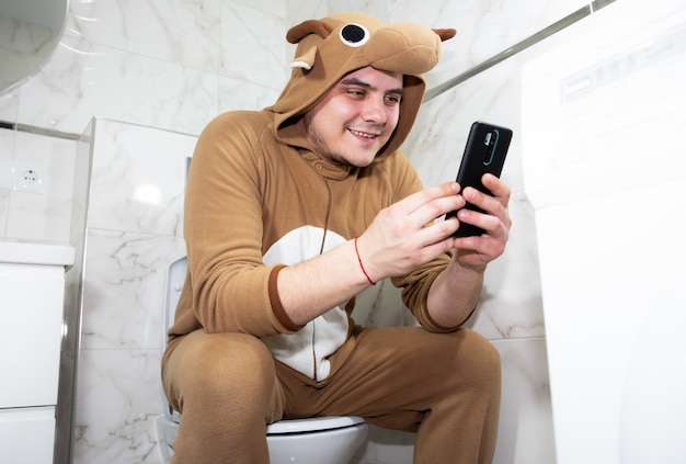 Man in cosplay costume of a cow. Guy in the funny animal pyjamas sit on the toilet and playing games on the mobile phone.