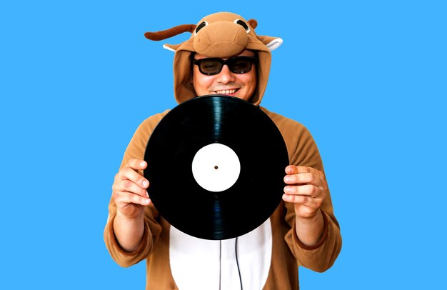 Man in cosplay costume of a cow with gramophone record isolated on blue background. Guy in the animal pyjamas sleepwear. Funny photo with party ideas. Disco retro music.