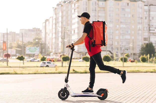 Человек-курьер с доставкой еды с красным тепловым рюкзаком едет по улице на электрическом скутере
