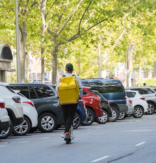 La consegna del cibo del corriere dell'uomo con lo zaino termico giallo percorre la strada su uno scooter elettrico consegna il cliente del cliente dell'ordine online