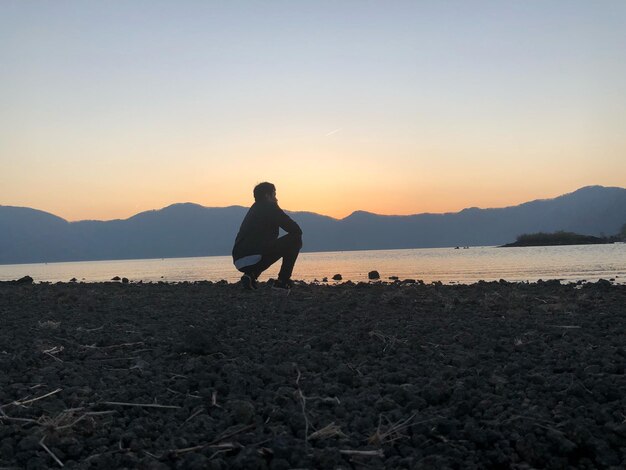 Foto uomo accovacciato contro il mare durante il tramonto