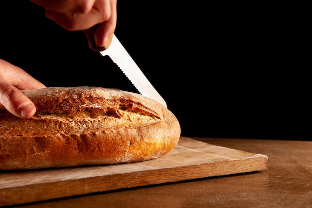 A man cuts baked bread with a knife on a black background Hands knife and bread