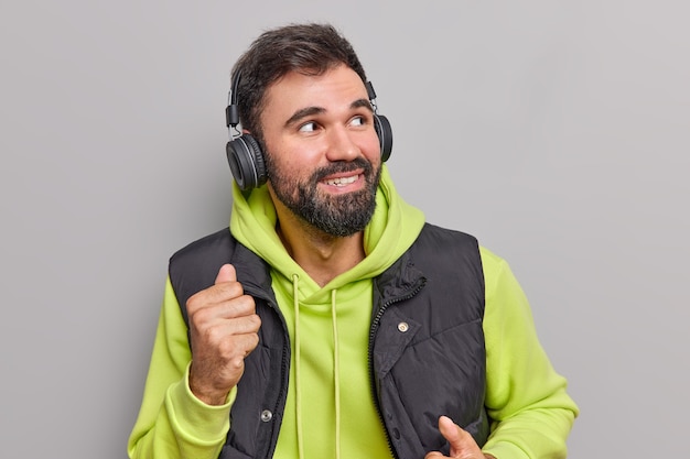 man dances carefree moves with rhythm of favorite music looks away wears wireless headphones on ears wears casual hoodie and vest isolated on grey