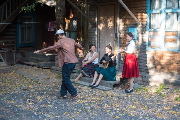 A man dancing russian folk dance women sitting on the stairs near the country house and talking to each other a woman playing balalaika