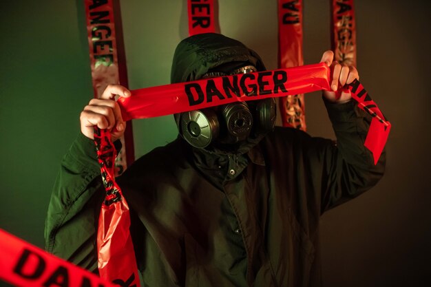 A man in a dark protective suit with a gas mask on his face and a hood on his head posing standing near a green wall holding danger tapes over his face. Danger concept