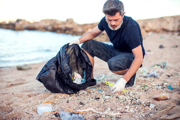 Uomo in camicia scura e pantaloncini con guanti bianchi e grande pacchetto nero che raccoglie immondizia sulla spiaggia. protezione ambientale e concetto di inquinamento del pianeta