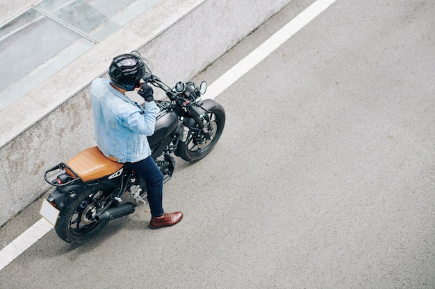 Photo man in denim jacket adjusting helmet and getting ready to ride on motorcycle