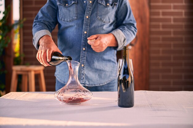 man in a denim shirt holding a bottle while pouring red wine into a glass decanter