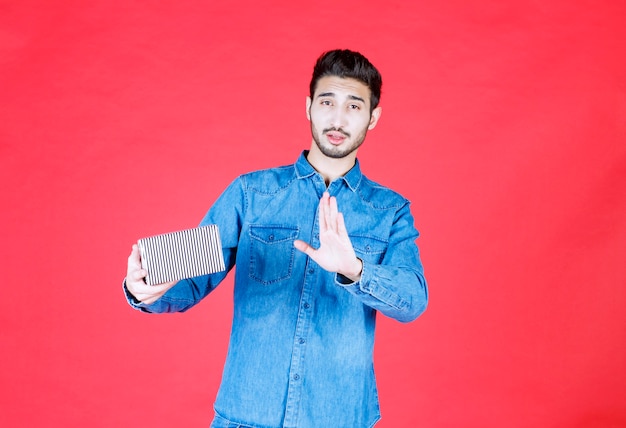 Man in denim shirt holding silver gift box and giving refusal.