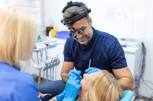 写真 男性歯科医と彼の助手は、ゴム手袋を着用して歯科用ツールを保持している患者の金髪女性の治療を行っています。口腔病学歯科現代の歯科医院のコンセプト