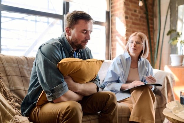 Man in depression sitting on sofa and listening to therapist during psycho session