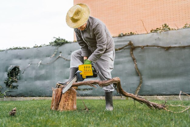 Man die een boomstam snijdt met een kettingzaag in de tuin, leunend op een andere boomstam op een zonnige dag