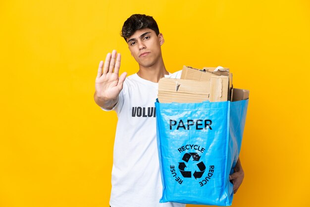 Man die een recyclingzak vol papier houdt om over geïsoleerde gele muur te recyclen en stopgebaar maakt