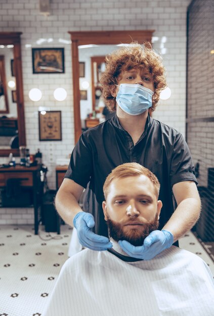 Man die haar laat knippen in de kapperszaak met een masker tijdens de pandemie van het coronavirus. Professionele kapper die handschoenen draagt. Covid-19, schoonheid, zelfzorg, stijl, gezondheidszorg en geneeskundeconcept.