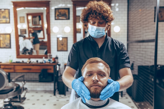 Man die haar laat knippen in de kapperszaak met een masker tijdens de pandemie van het coronavirus. Professionele kapper die handschoenen draagt. Covid-19, schoonheid, zelfzorg, stijl, gezondheidszorg en geneeskundeconcept.