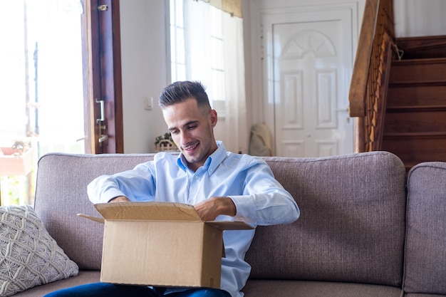 Man die het pakket thuis uitpakt. Gelukkig jonge man kijken naar kartonnen doos zittend op de bank in de woonkamer thuis. Man die thuis afgeleverde spullen afrekent.